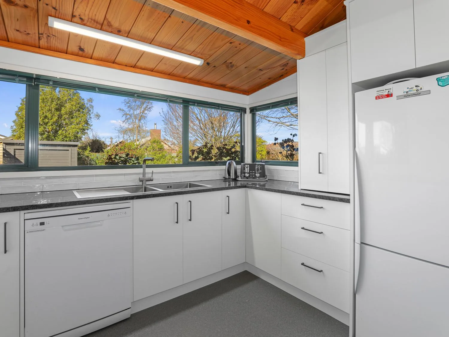 Una cocina luminosa tiene armarios blancos, encimeras oscuras y amplias ventanas con vistas a los árboles.