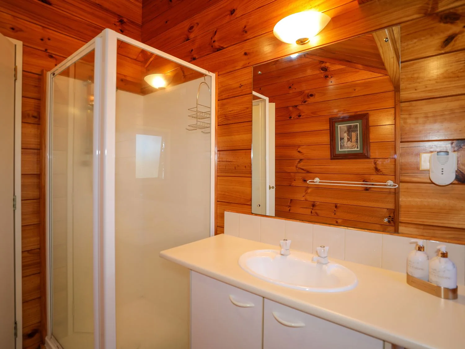 Un baño con ducha y lavabo bajo un espejo grande, rodeado de paredes revestidas de madera.