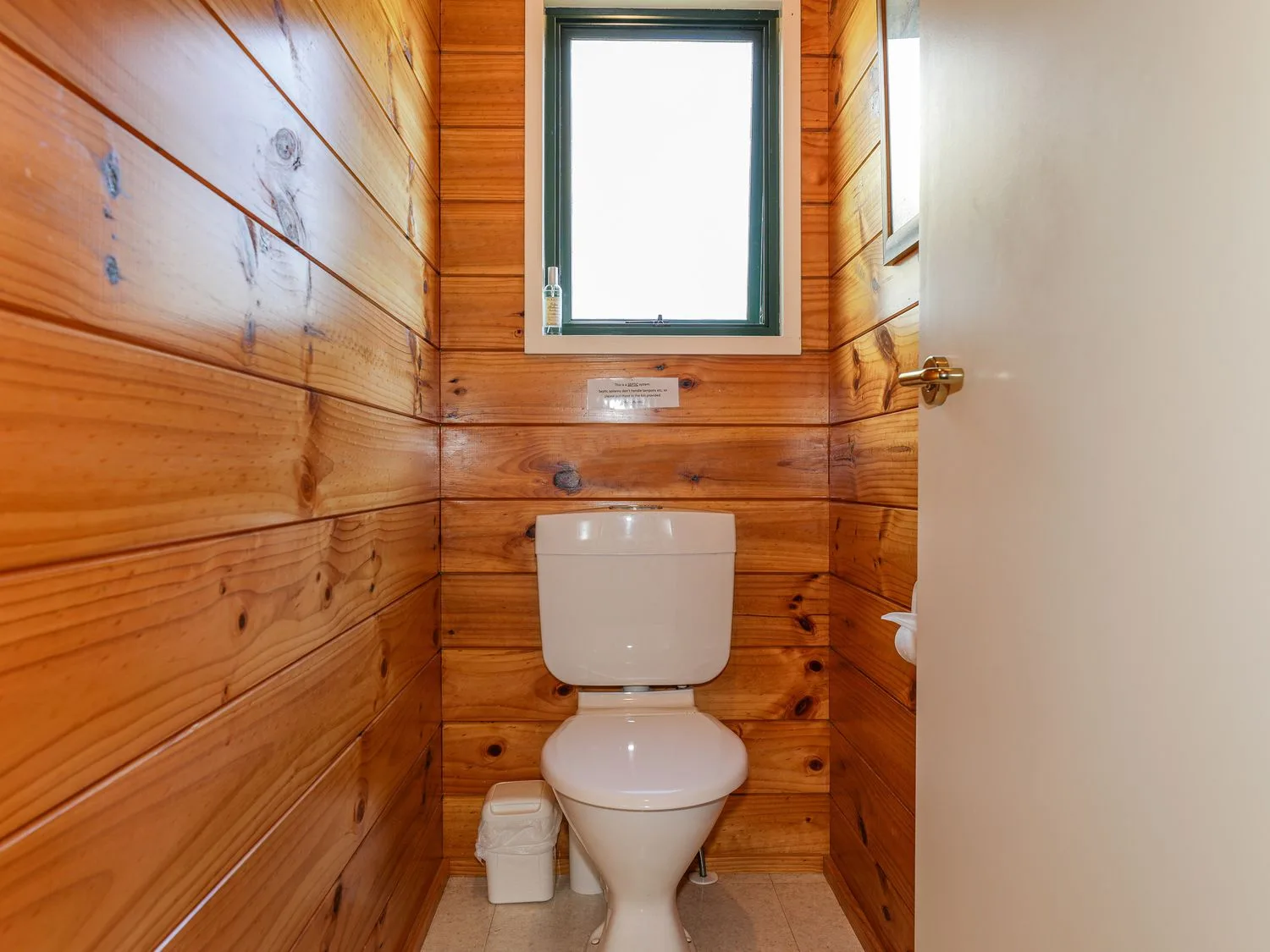 Un baño compacto con un inodoro blanco, paredes revestidas de madera y una ventana pequeña.