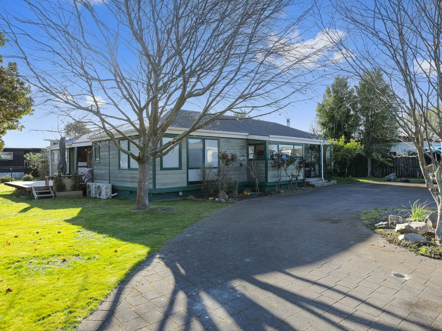 Una casa de una sola planta con molduras verdes, junto a una entrada pavimentada para vehículos, con árboles sin hojas y un jardín con césped en Five Mile Yay - Five Mile Bay Holiday Home, en Waitahanui, a 12 km de Taupo.