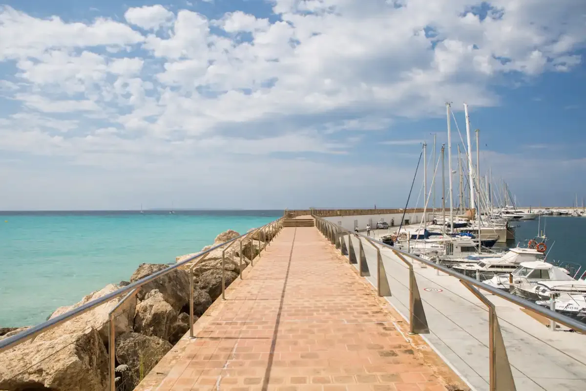 A seaside marina walkway extends between turquoise water and moored sailboats.