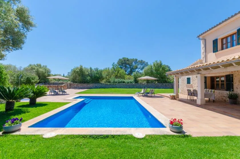A swimming pool sits beside a tiled patio and a house, with a lawn in front.