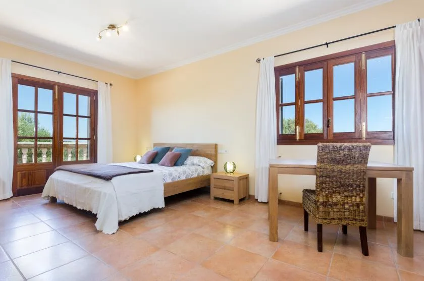 A bedroom with a bed and a desk beside large windows.