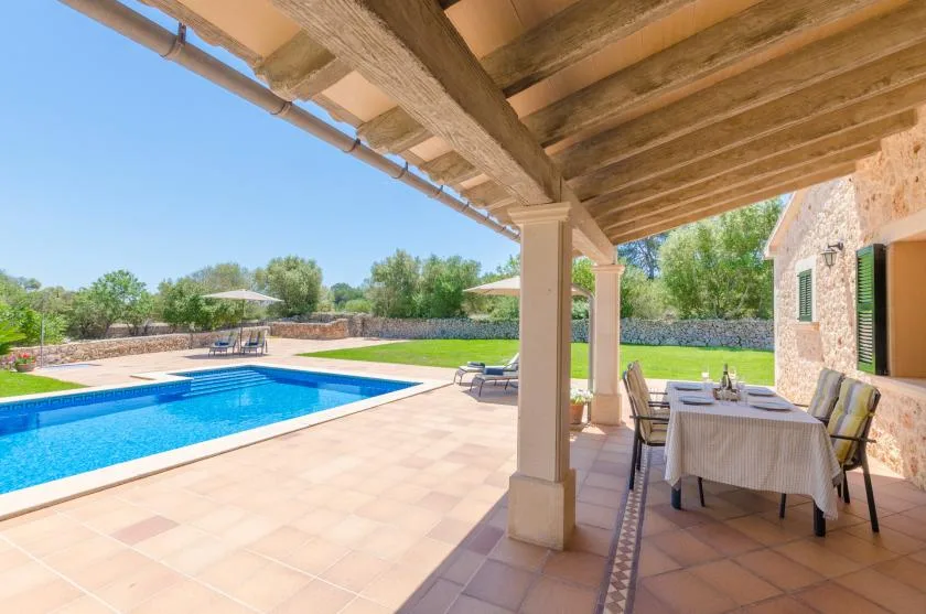 A covered patio with outdoor dining overlooks a swimming pool and garden.