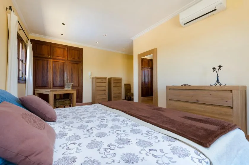A bedroom with a patterned bedspread, wooden furnishings, and an air conditioner.
