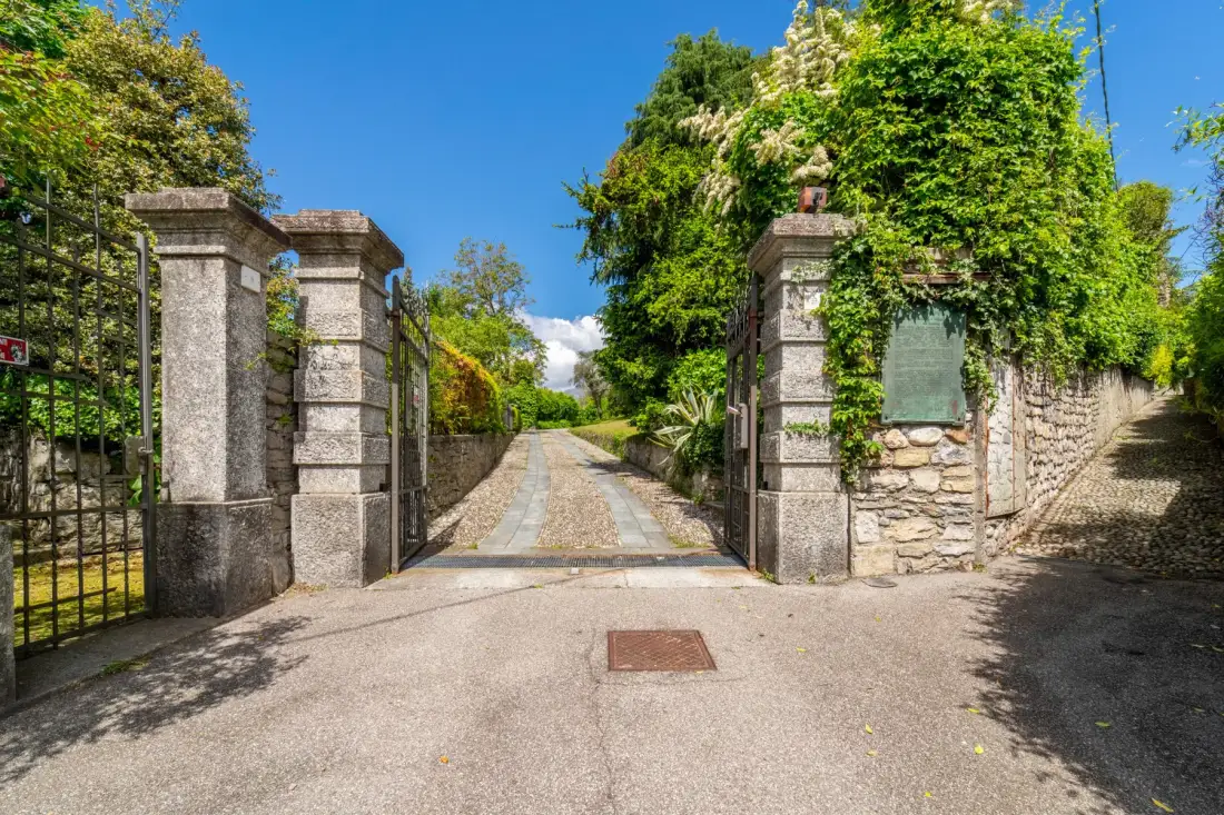 A stone entrance opens onto a paved lane bordered by greenery.