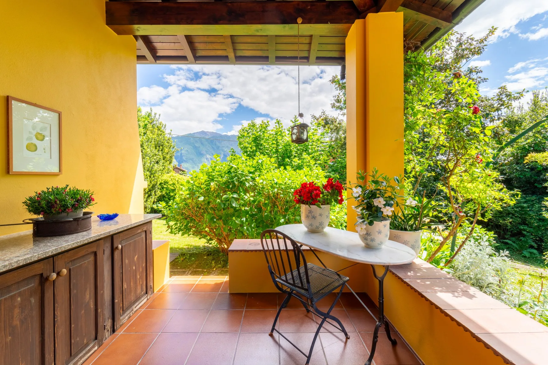 A covered patio with a table and chairs overlooks a garden and distant hills.