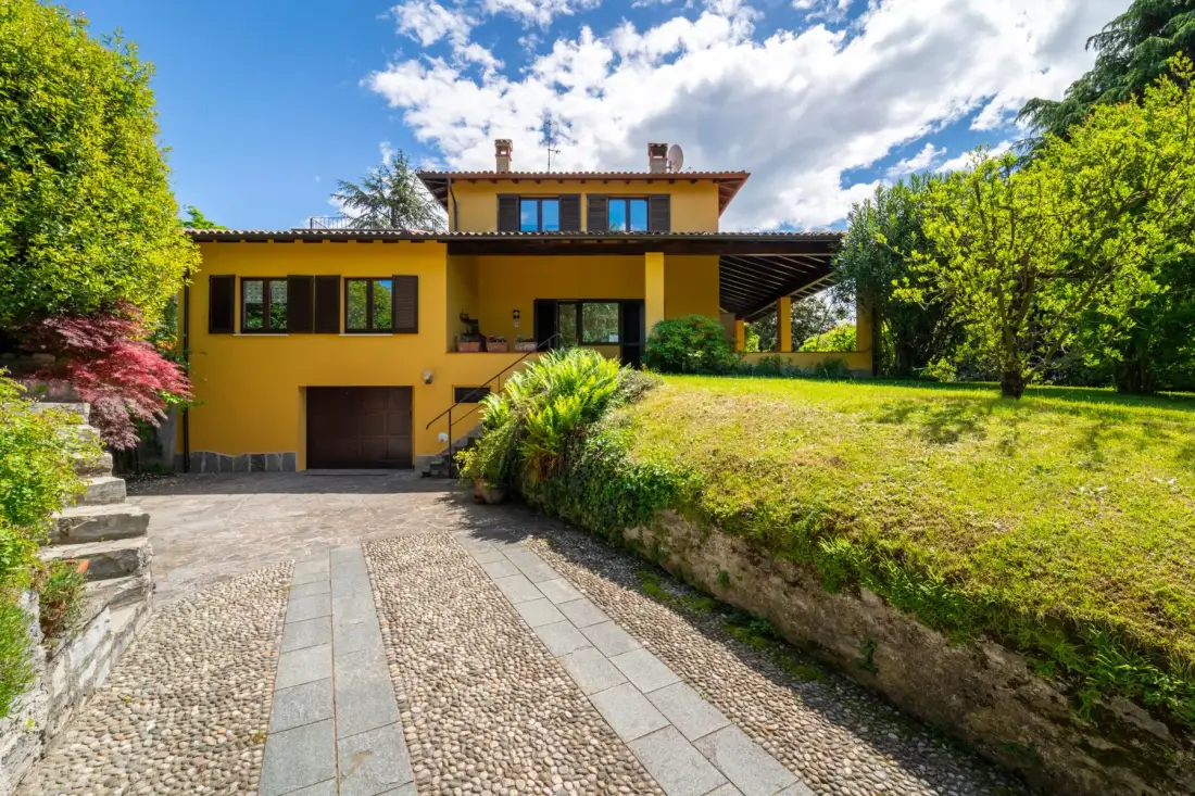 A yellow house stands beside a sloping lawn, with a paved driveway in front.