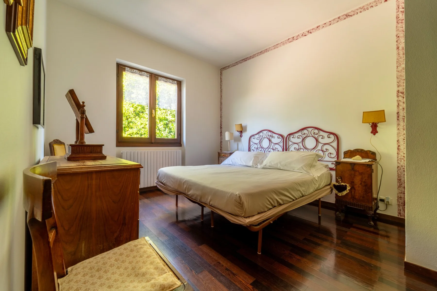 A bedroom with a large bed, wooden furnishings, and a window overlooking greenery.