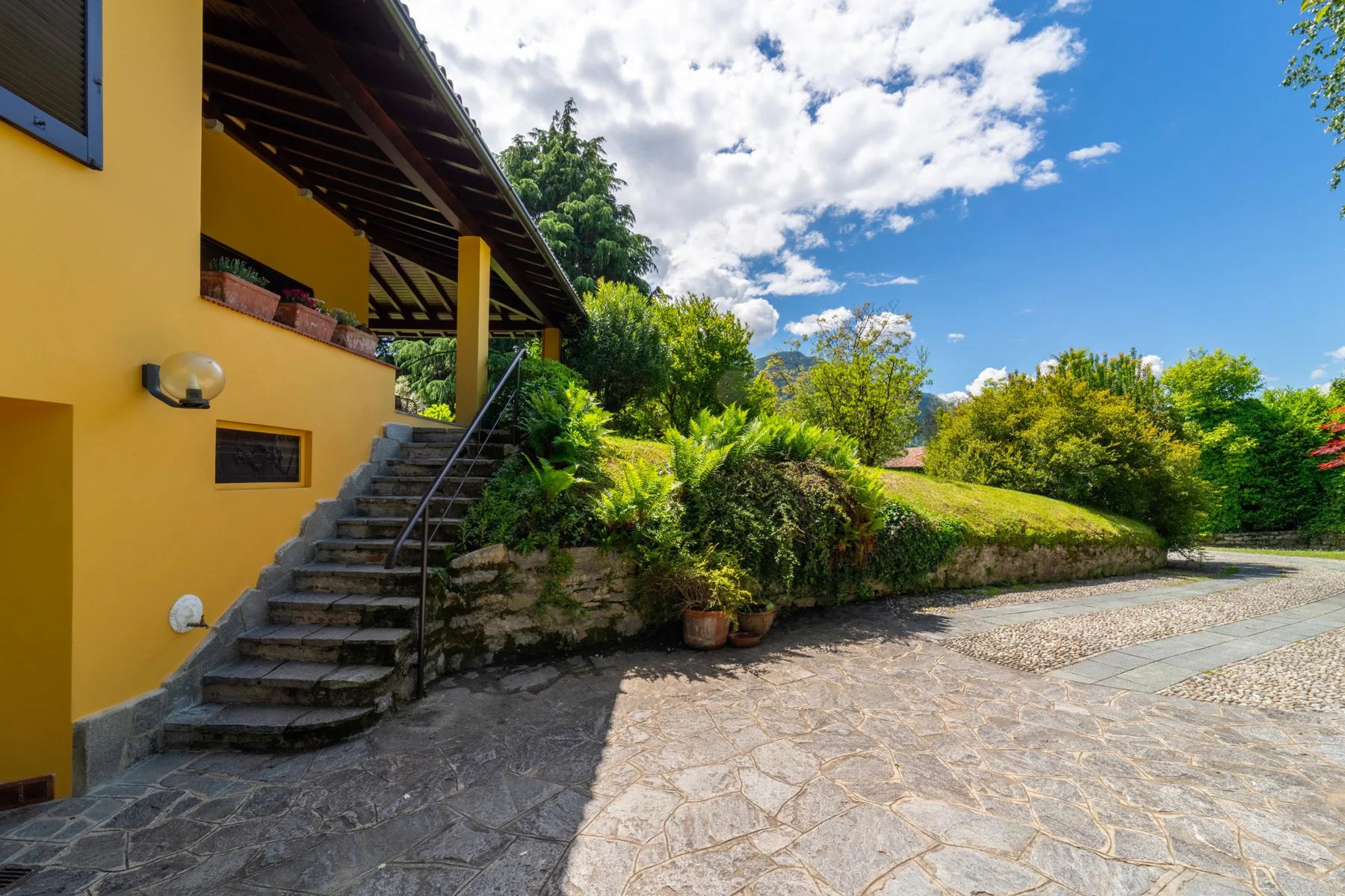 A yellow building beside a stone-paved courtyard, with steps and lush greenery.