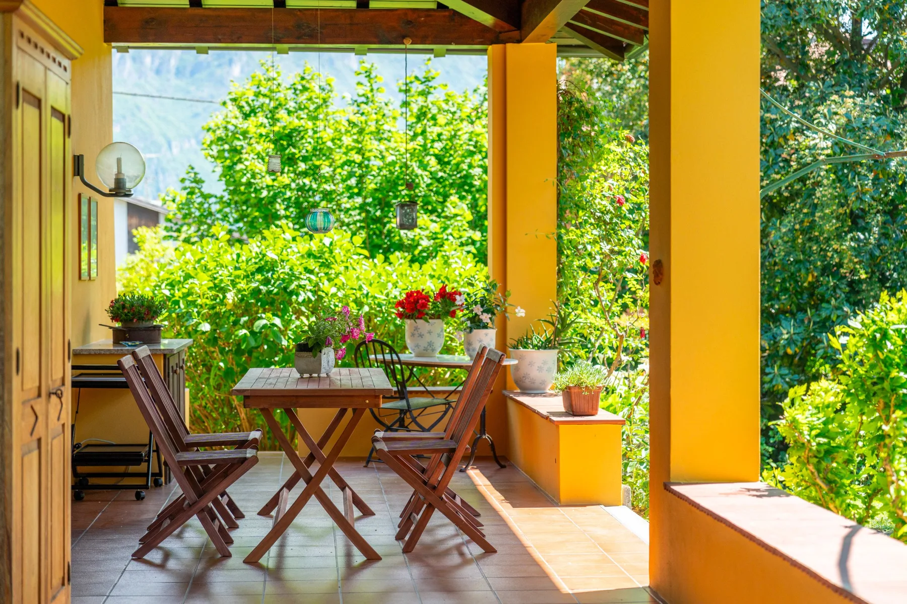 A covered patio has a wooden table and seating surrounded by lush greenery.