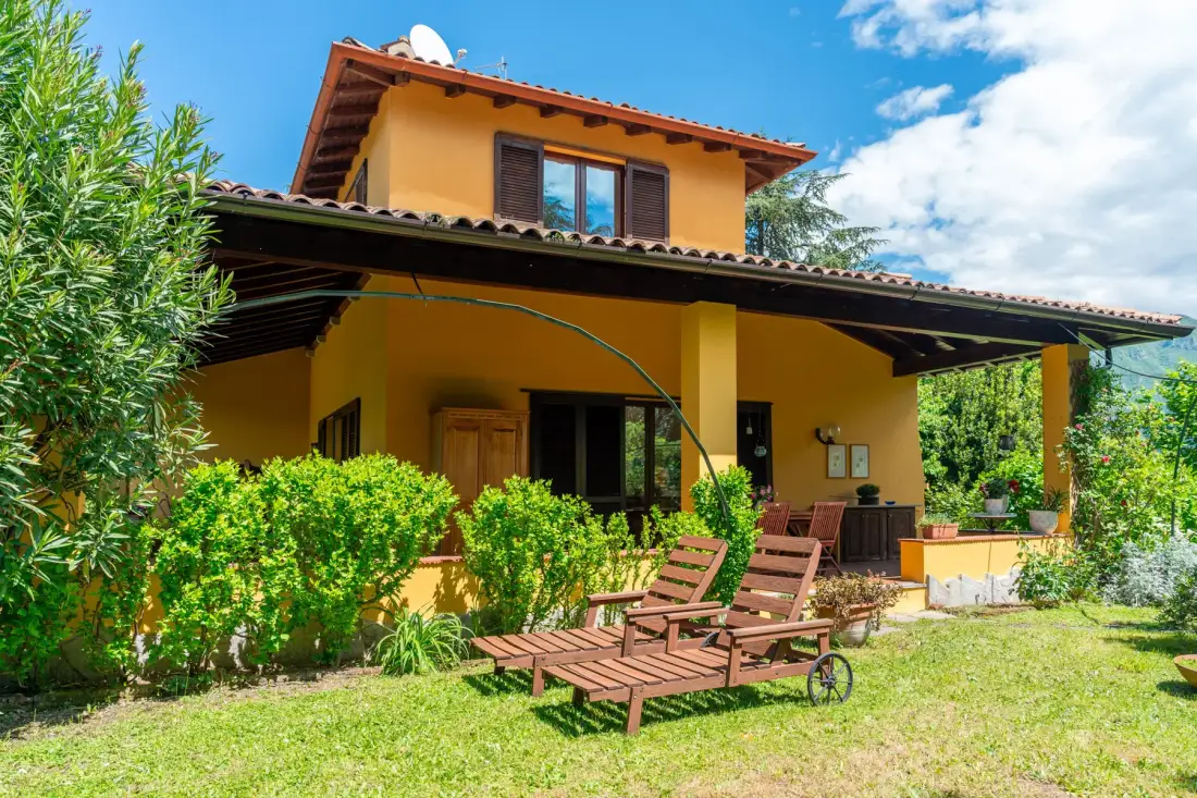A yellow house with a covered patio and wooden seating in the garden.