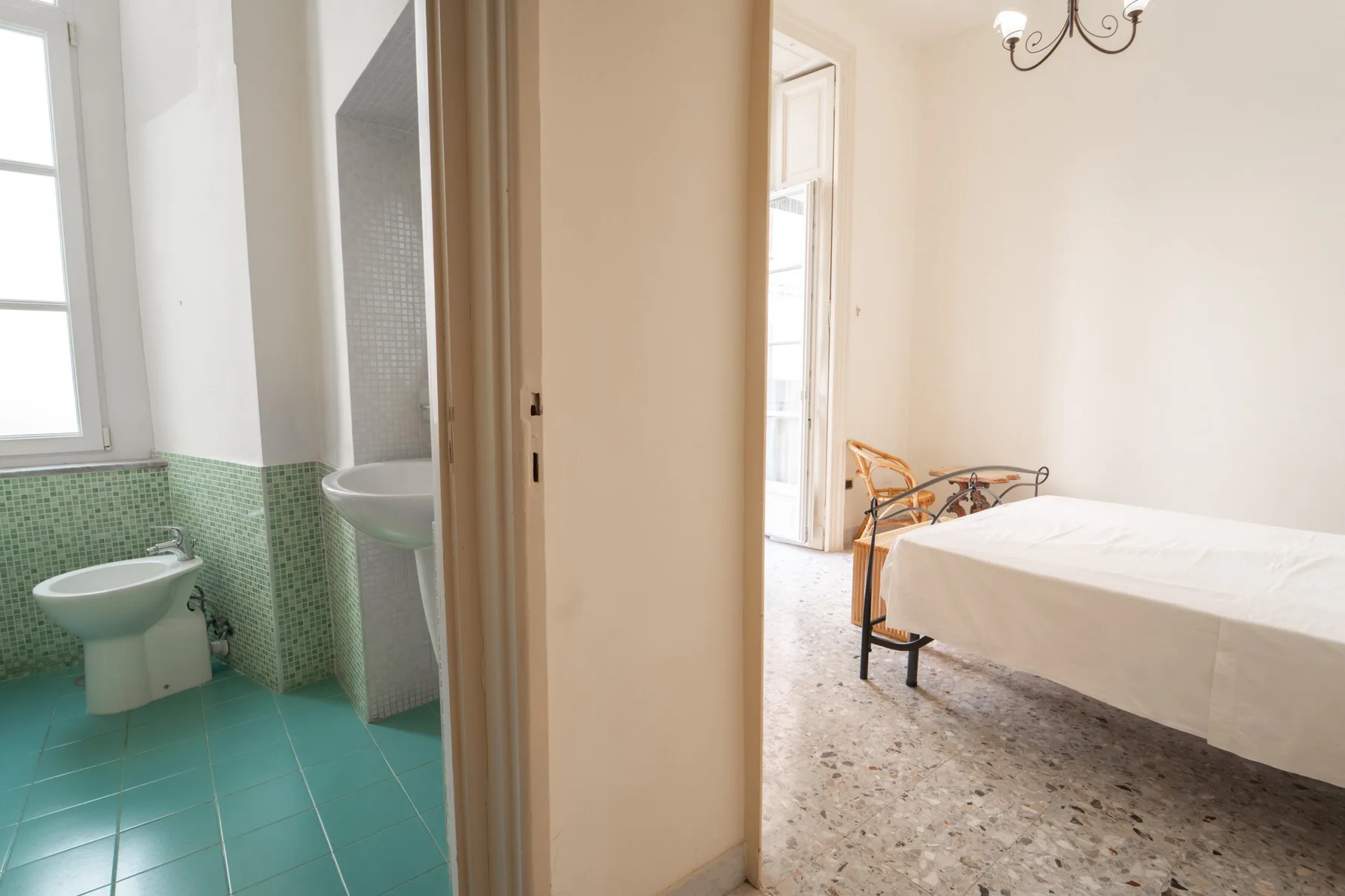 A bright bedroom with a white bed beside an open doorway to a tiled bathroom.