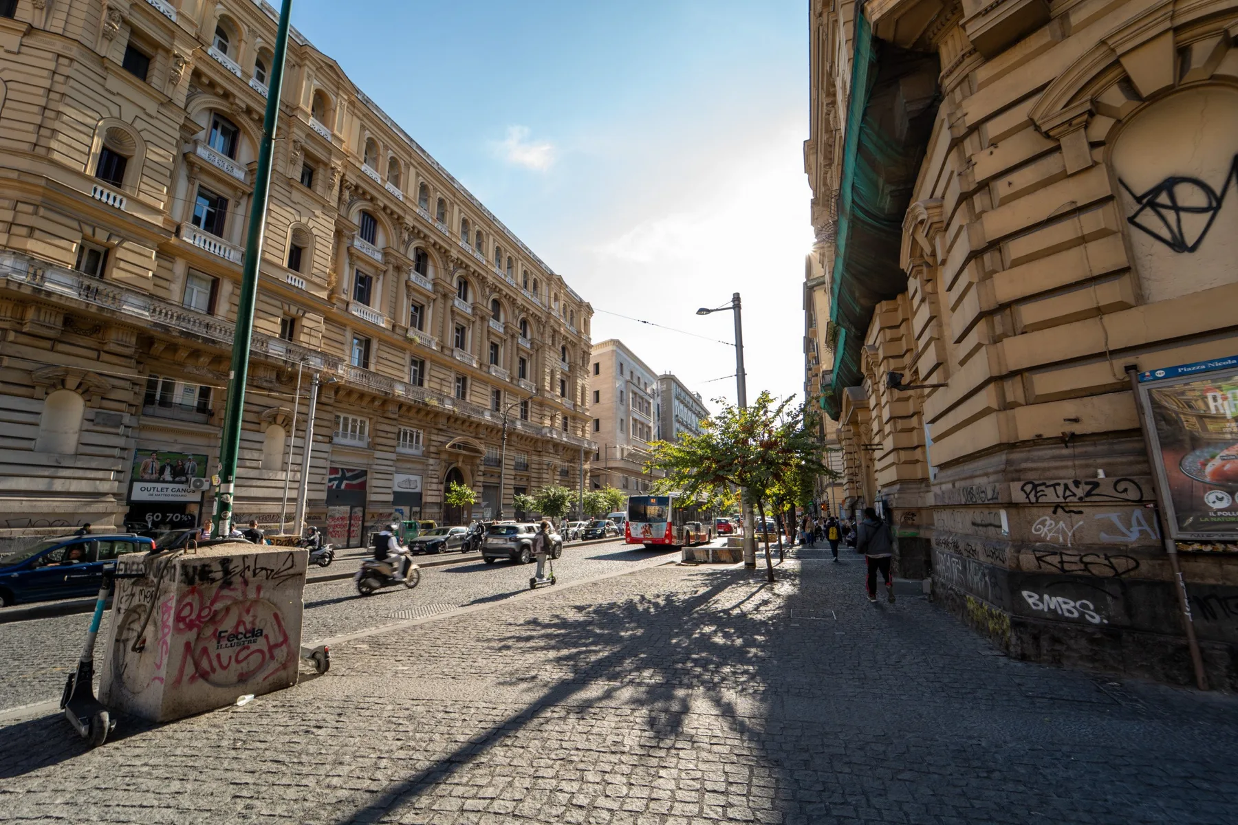 A cobblestone city street lined with tall stone buildings and parked vehicles.
