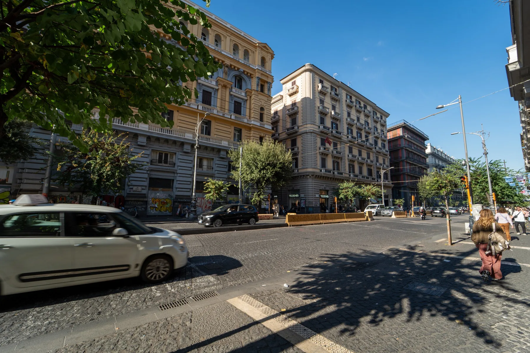 An urban street with multi-story buildings, trees, and passing traffic.