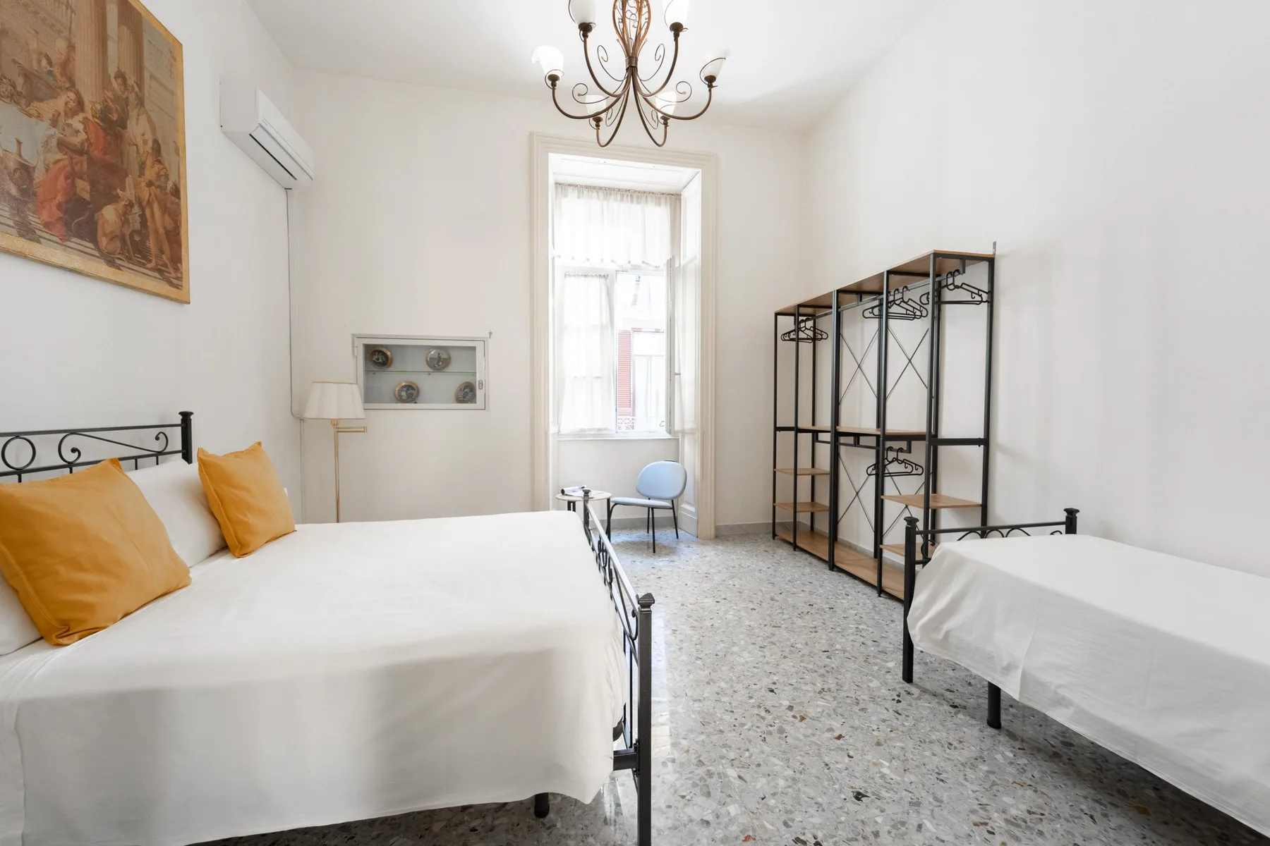 A bright bedroom with two beds, a central window, and open shelving.