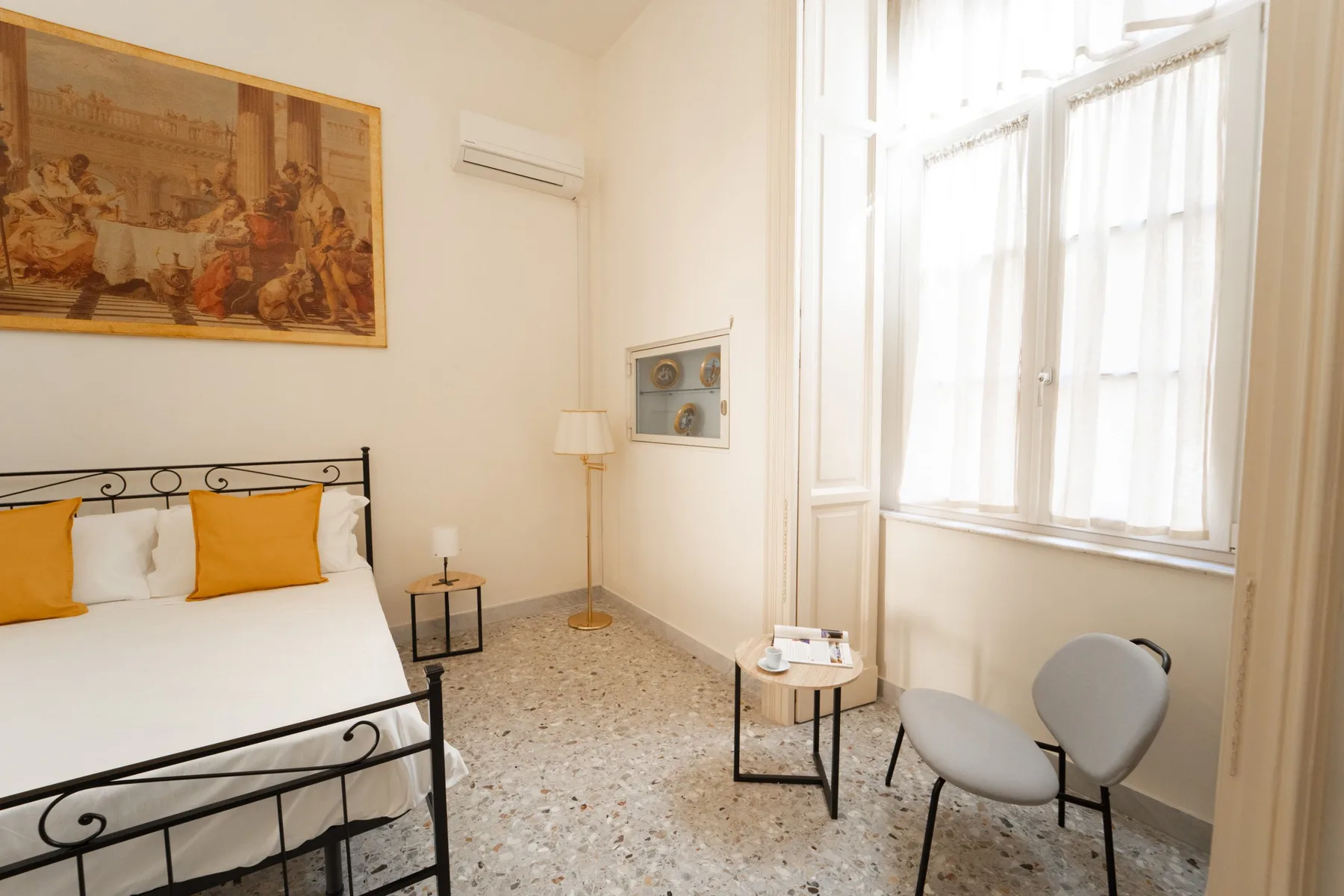 A bright bedroom with a bed, a large curtained window, and a small seating area.