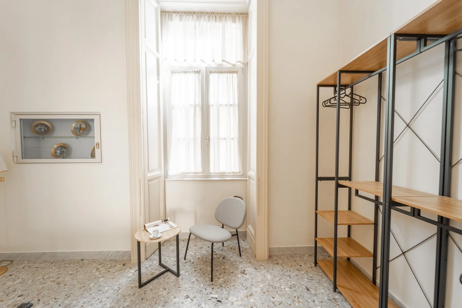 A bright room with tall curtained windows, a small table, and open shelving.
