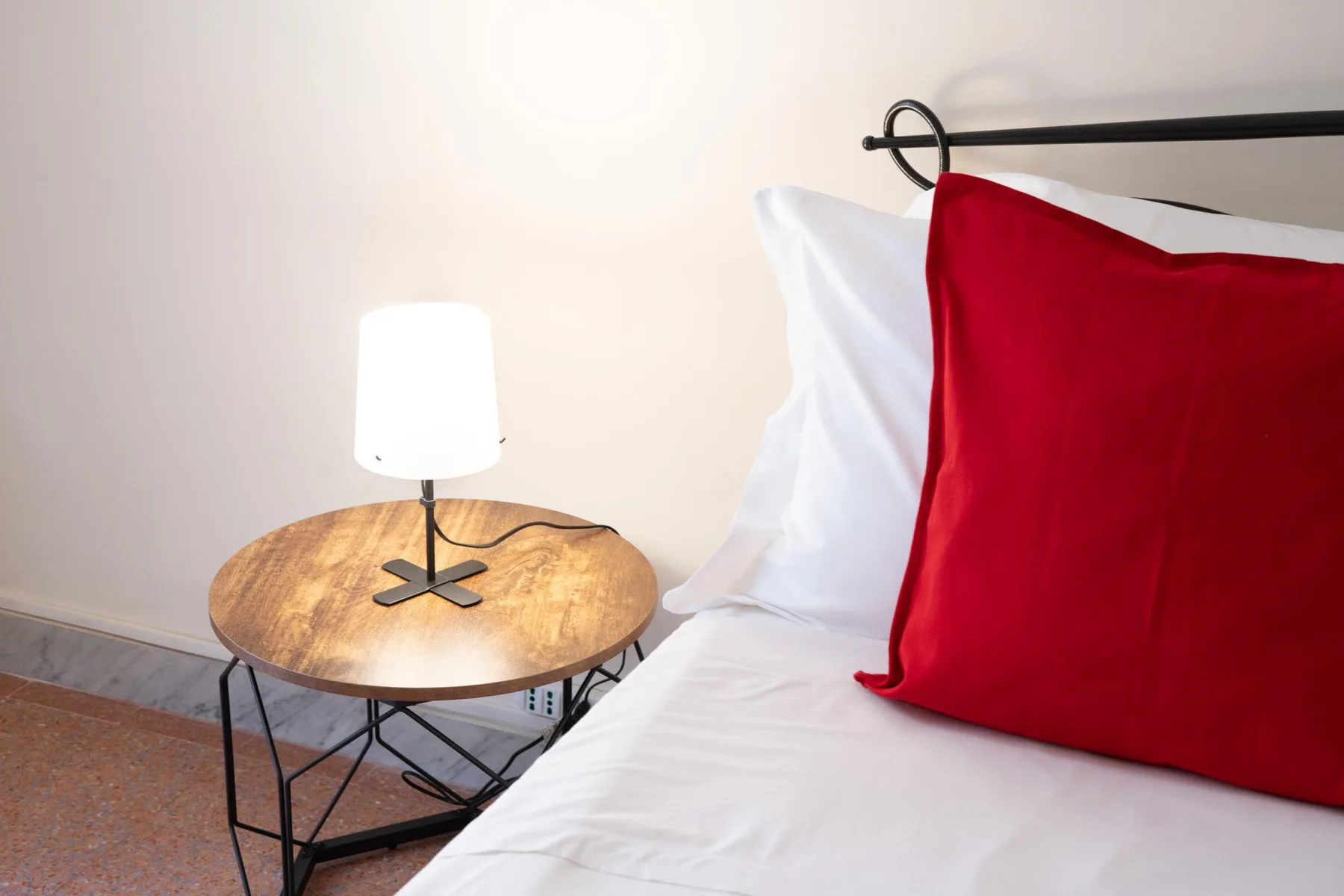A bedroom with white bedding and a red pillow beside a small table lamp.