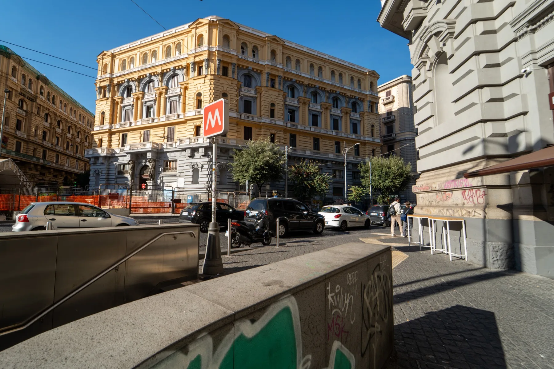 A city street with a metro entrance, parked cars, and ornate buildings.