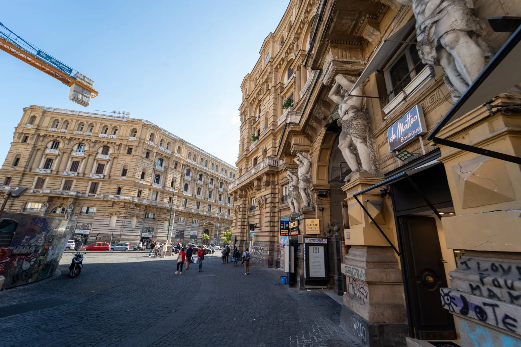 A city street lined with ornate historic buildings, with pedestrians walking along the pavement.