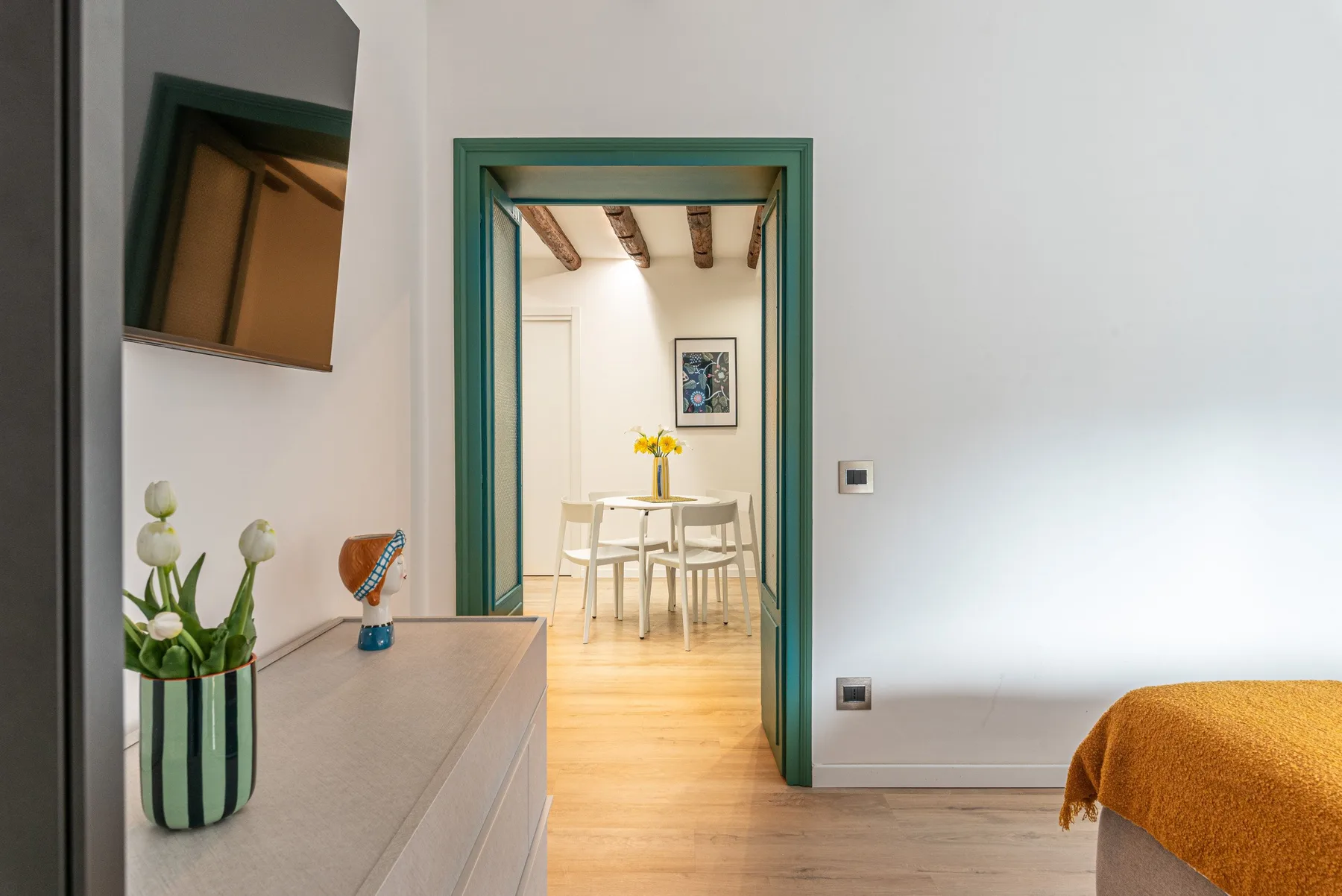 Un dormitorio se abre, a través de una puerta con marco verde, a un comedor con mesa y sillas.