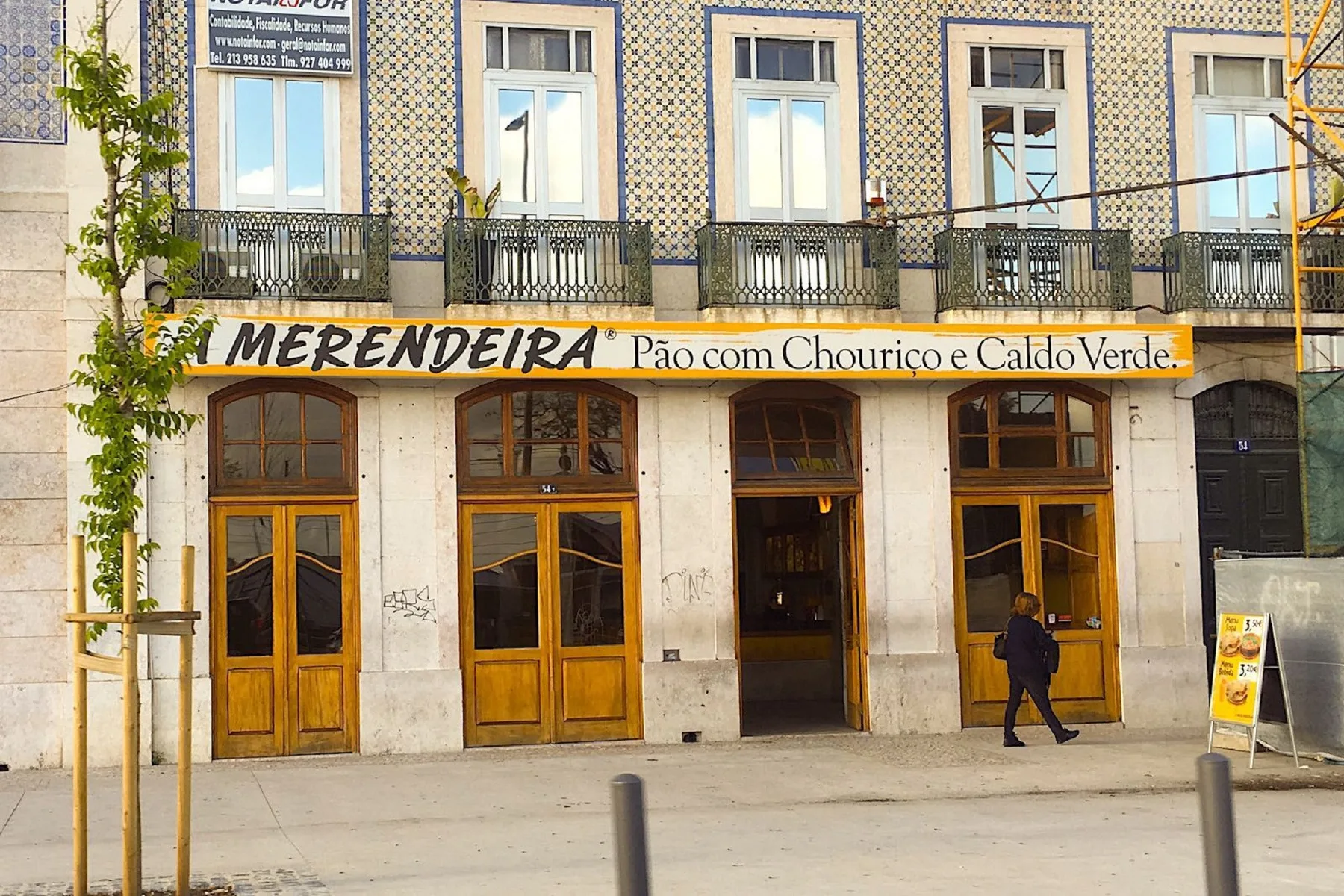 Una cafetería a pie de calle bajo una fachada de azulejos, con puertas y ventanas de madera.