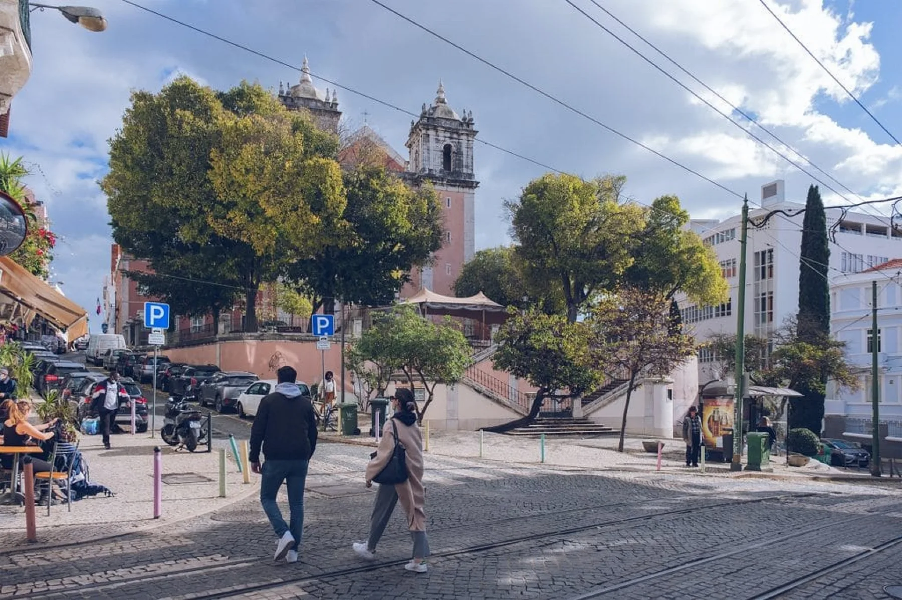 Una calle urbana con peatones, coches estacionados, árboles y una torre de iglesia.