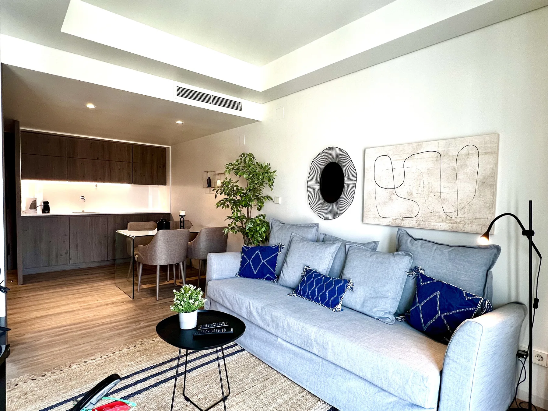 Una sala de estar con un sofá claro, cojines azules y una pequeña zona de comedor.
