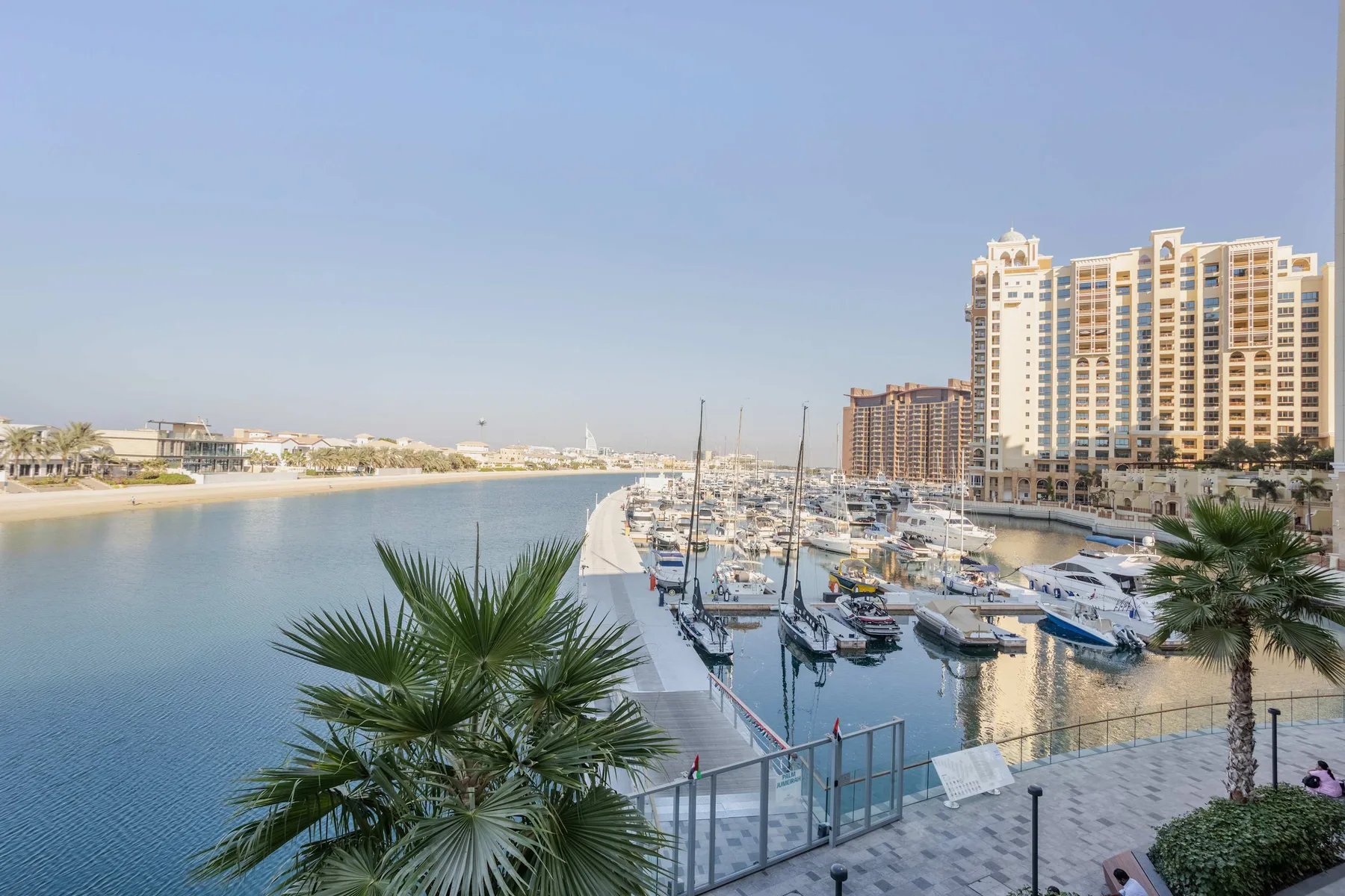 A marina lined with boats sits beside waterfront apartment buildings and palm trees.