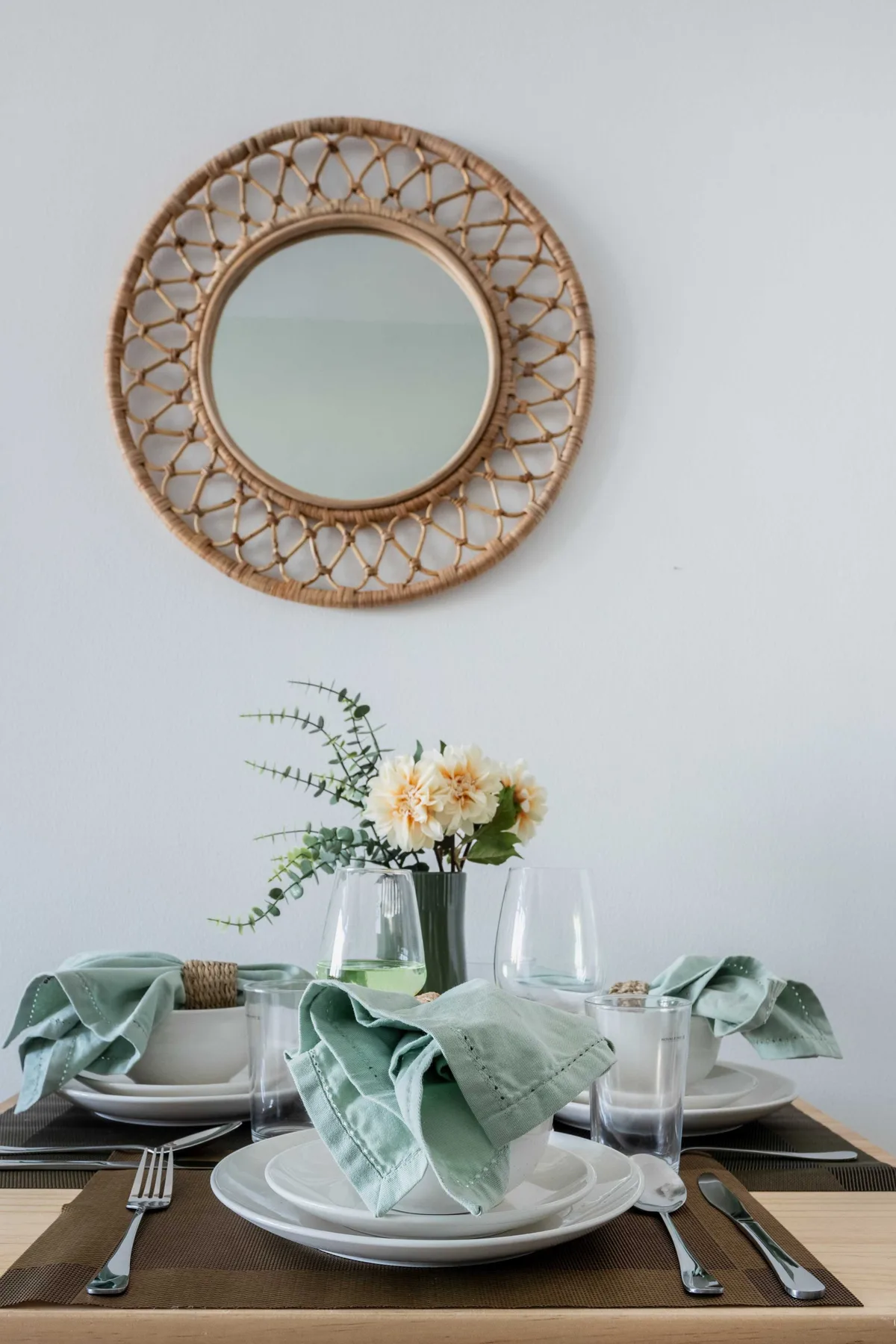 A dining table is set with plates and glasses beneath a round woven wall mirror.