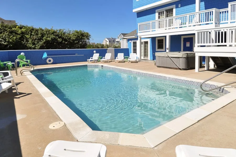 Una piscina está junto a una casa azul con balcones blancos y asientos alrededor.
