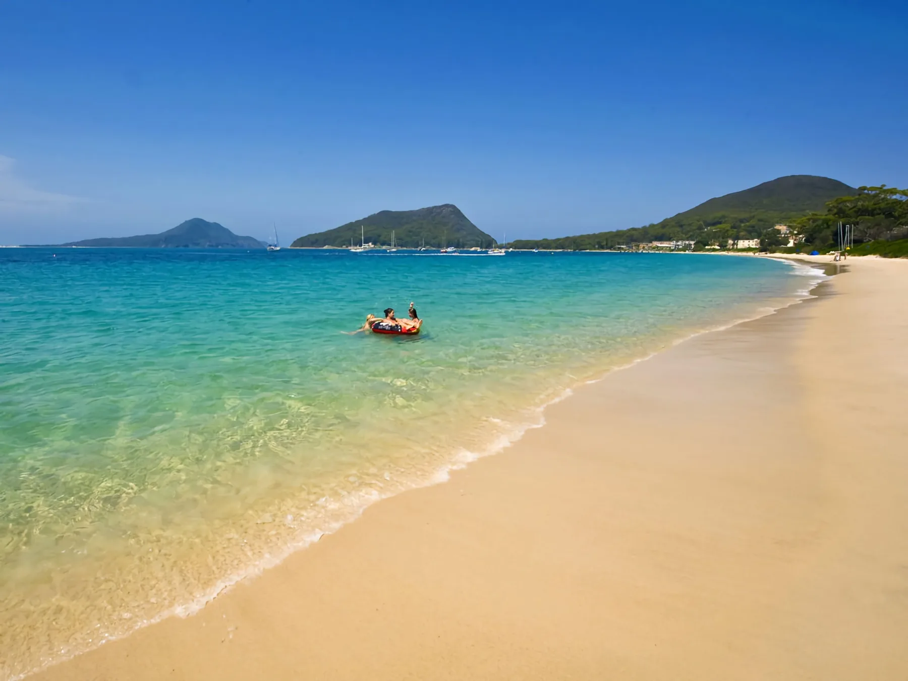 A sandy beach meets clear turquoise water, with hills across the bay.