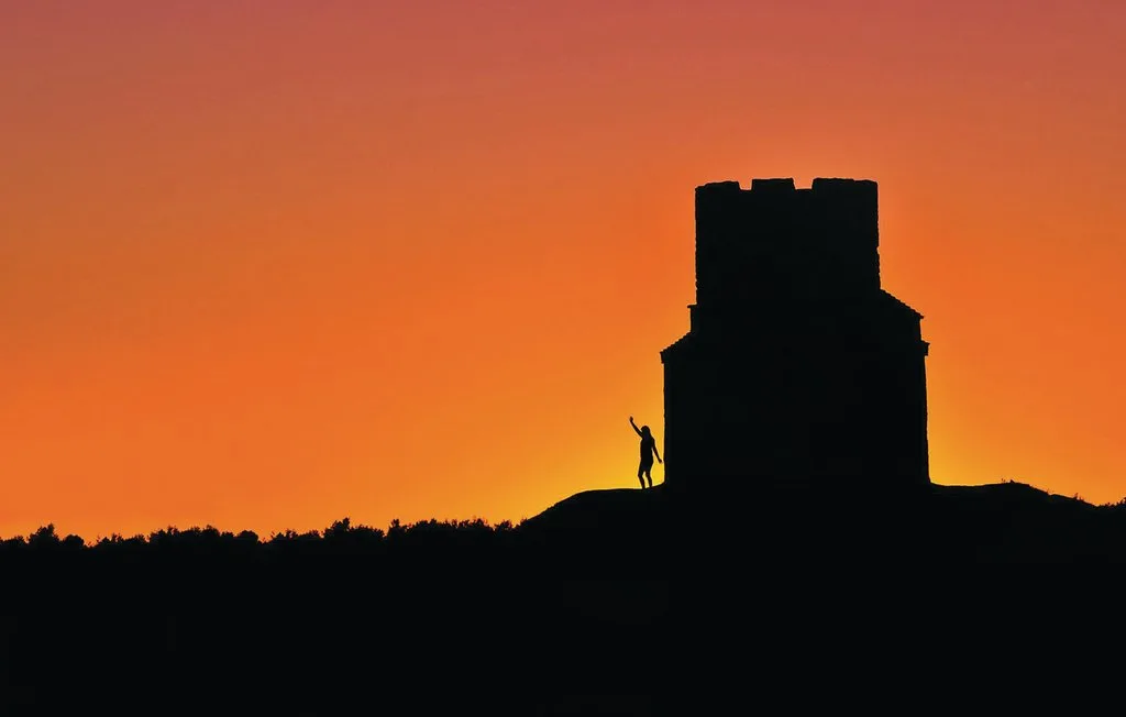A silhouetted tower stands on a ridge against a glowing orange sunset.