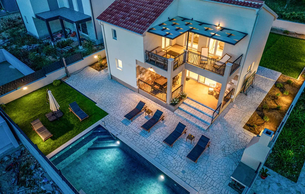 An illuminated two-story house overlooks a swimming pool and paved patio.