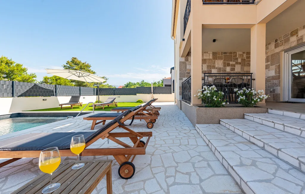 A paved poolside patio has wooden loungers, umbrellas, and a planted terrace beside the building.