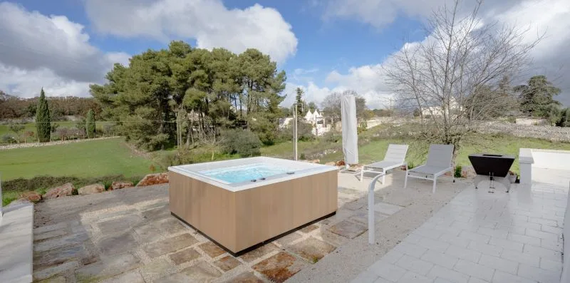 A paved outdoor patio features a raised hot tub, lounge chairs, and a garden view.