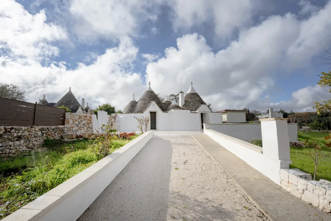 A stone driveway leads toward white buildings with conical roofs under a cloudy sky.