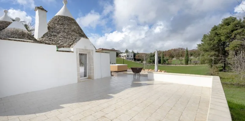 A paved outdoor terrace beside white buildings, with a lawn and trees beyond.