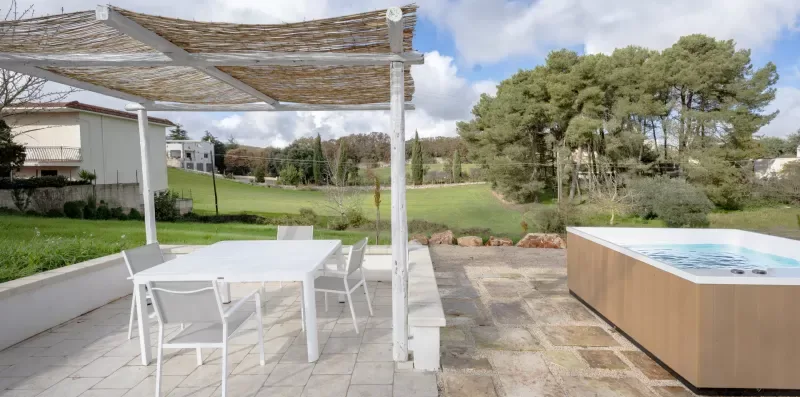 An outdoor patio has a dining table beneath a slatted canopy beside a hot tub.