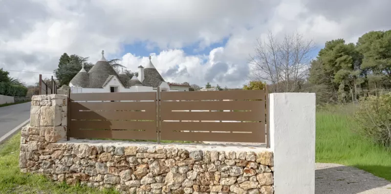 A brown entrance gate stands between stone walls beside a grassy roadside.