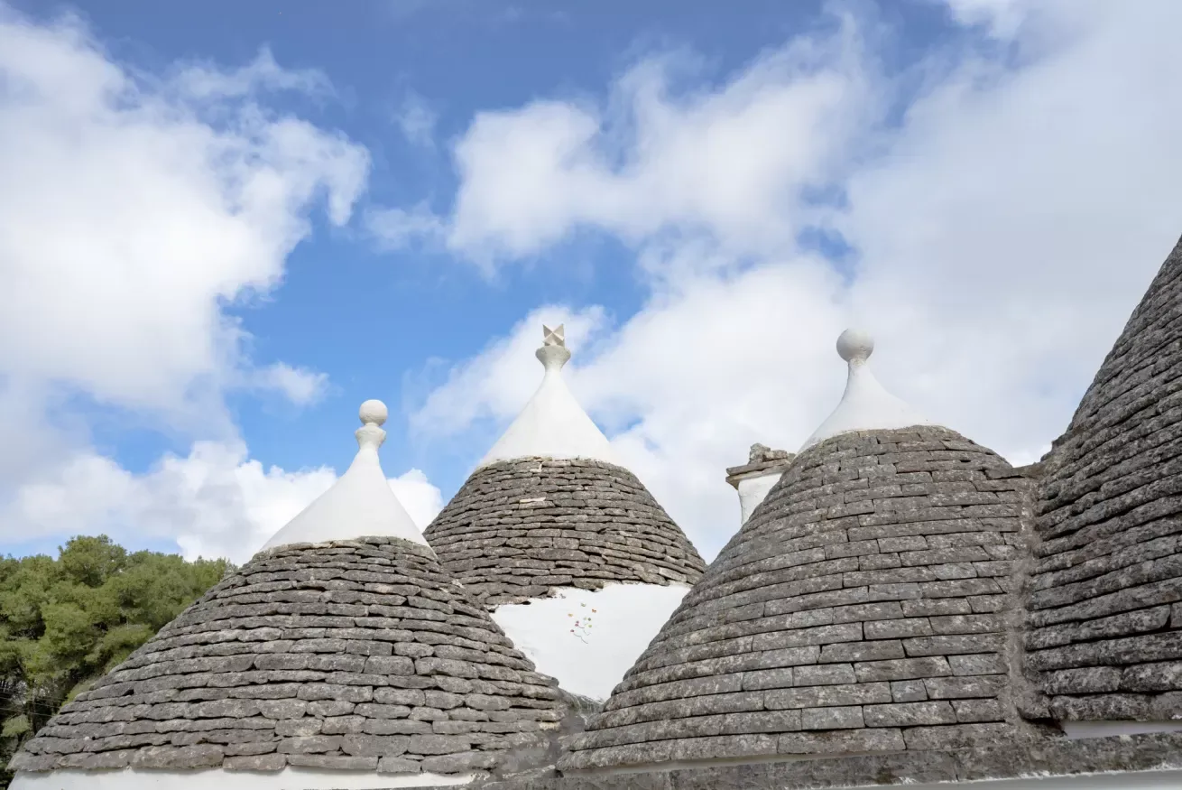 Stone-roofed trulli buildings with conical tops stand beneath a partly cloudy sky.