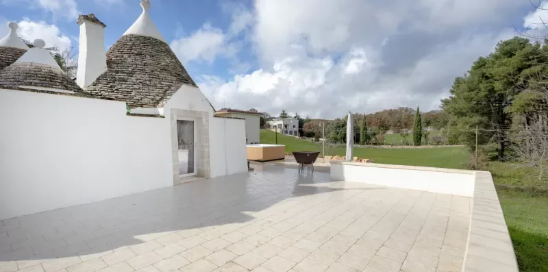 A paved outdoor terrace beside a white building, with a lawn and trees beyond.