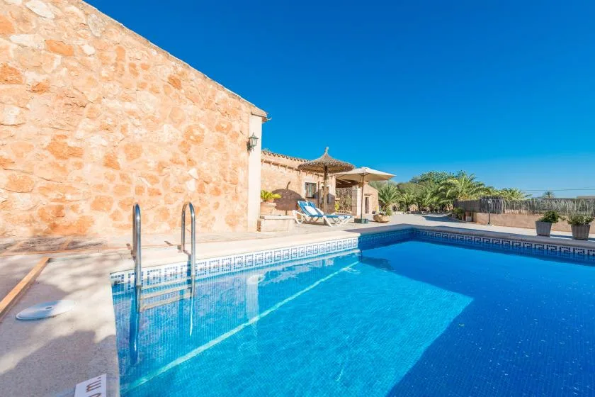 A swimming pool beside a stone building, with a shaded seating area in the background.