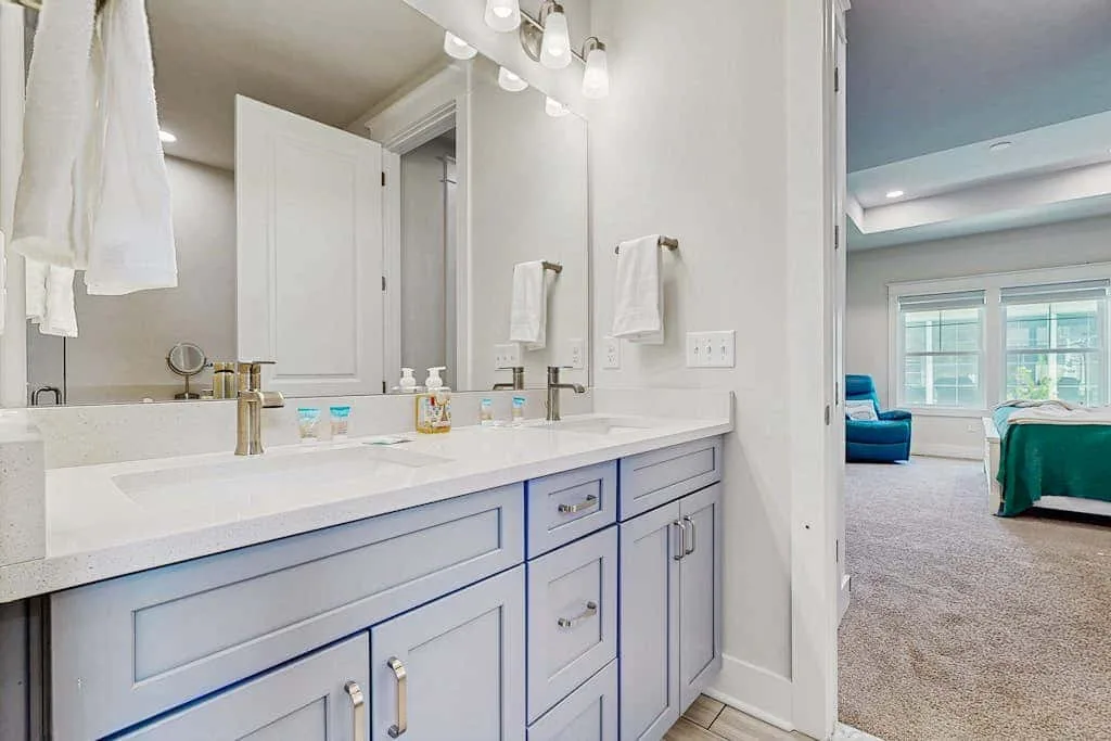 A bright bathroom vanity with a long sink counter opens onto a bedroom.