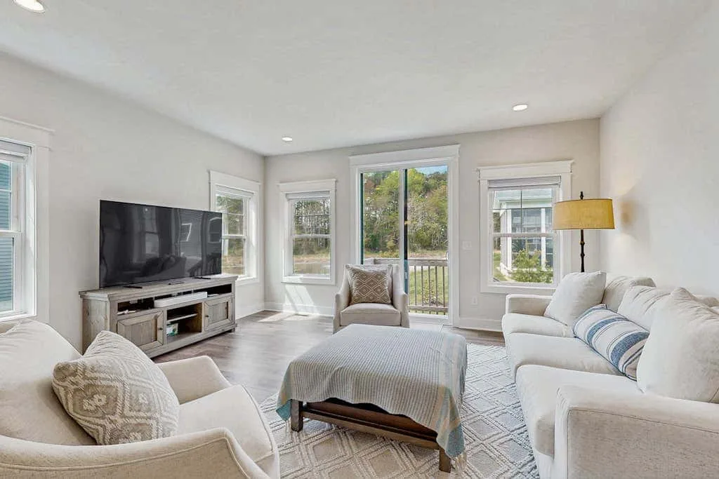 A bright living room with a television, seating, and windows overlooking greenery.