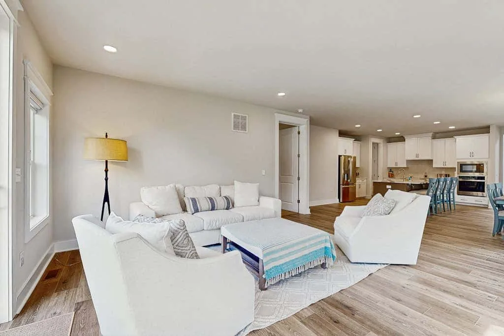 A bright open-plan living room with white seating and a kitchen beyond.