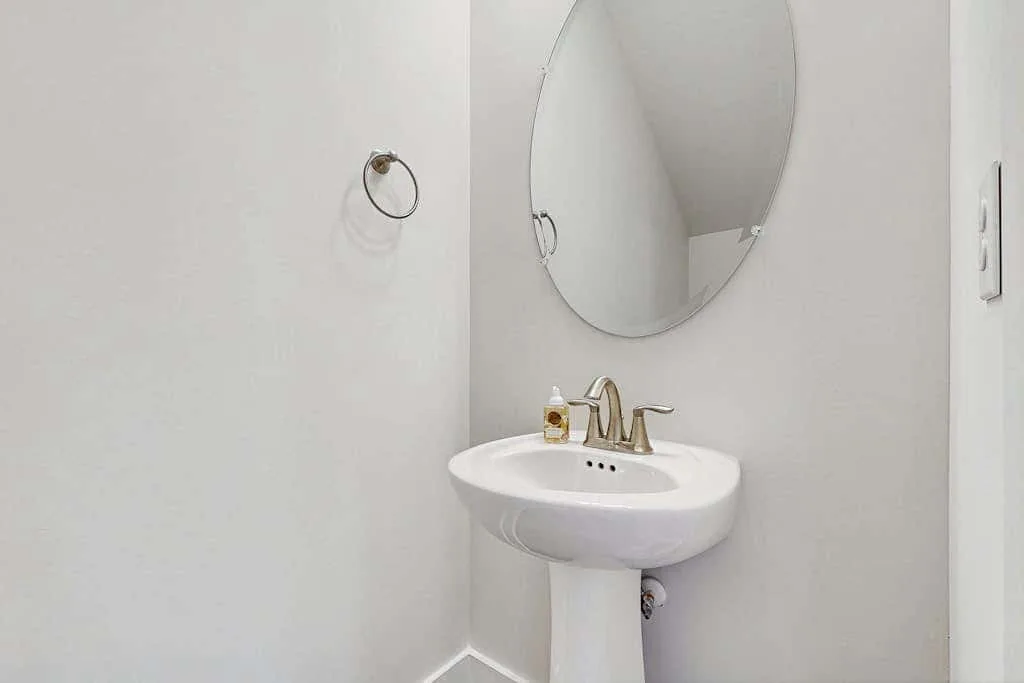A bathroom sink sits beneath a round mirror, with a towel ring on the wall.