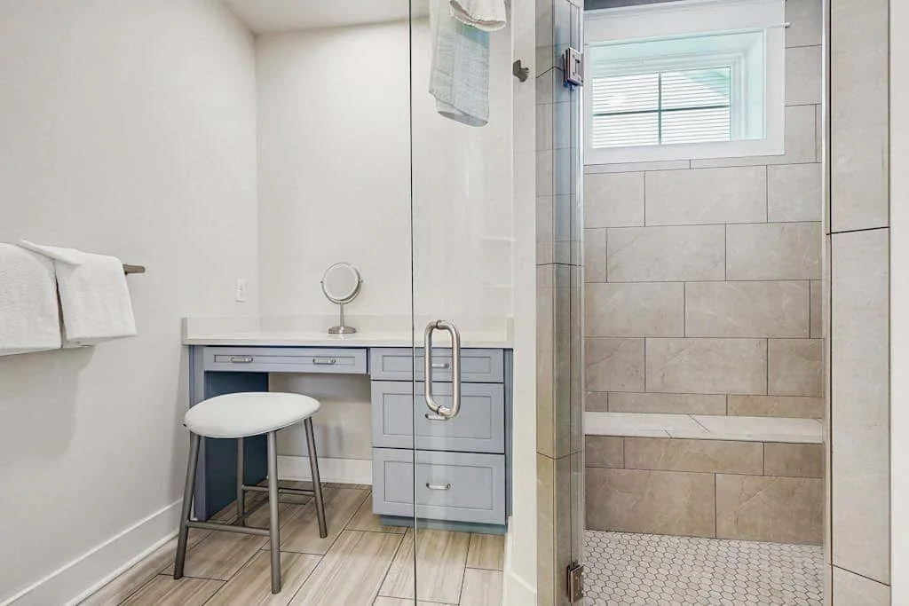 A bathroom vanity with a stool sits beside a glass shower and window.