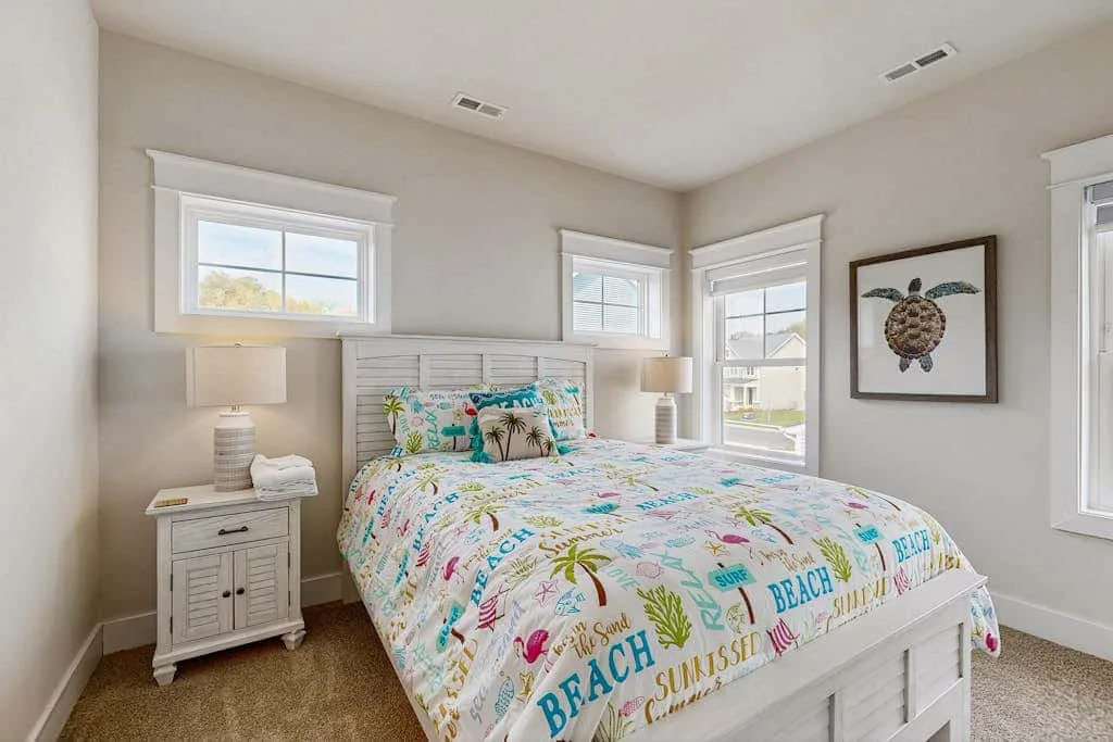 A bright bedroom features a bed with colorful beach-themed bedding and several windows.